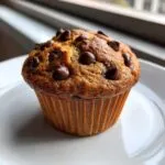 A close-up of a freshly baked chocolate chip muffin with melted chips on top, sitting on a white plate.