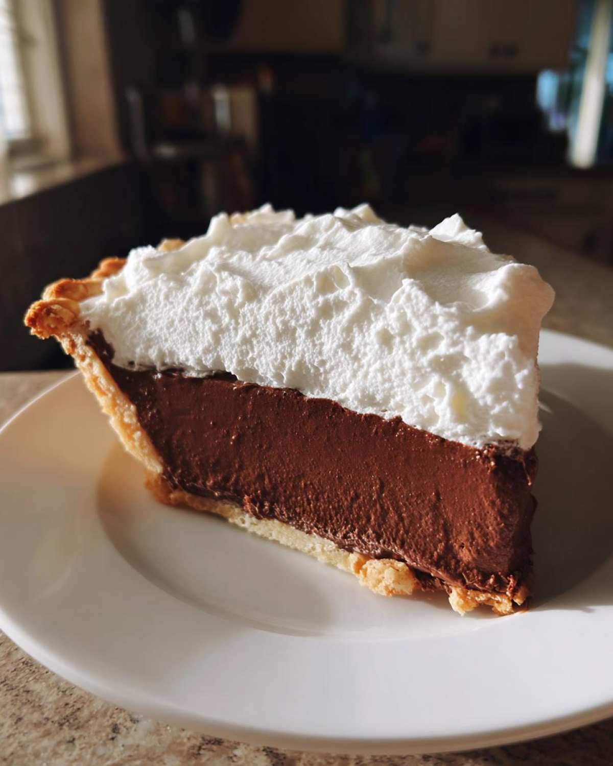 A perfect slice of chocolate cream pie with a thick chocolate layer and high mound of whipped cream on a white plate.