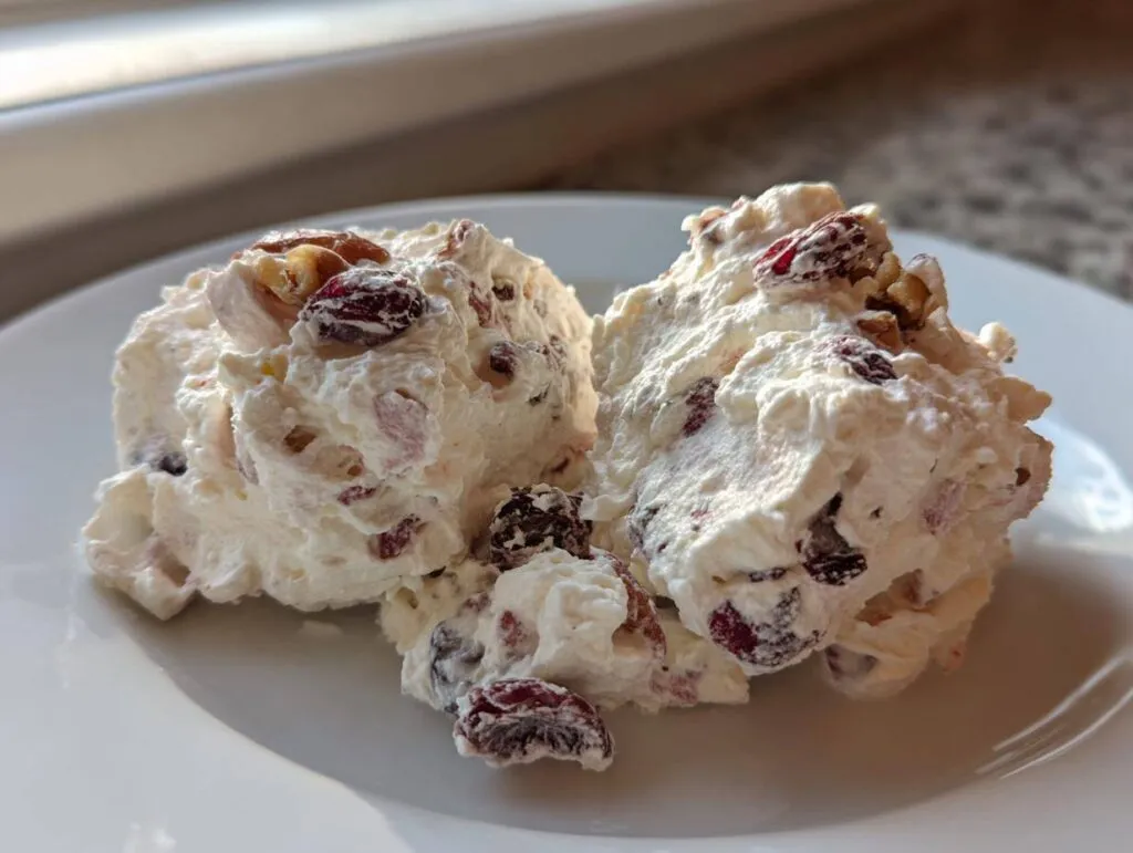 Two scoops of creamy, white cranberry salad mixed with dried cranberries and walnuts on a white plate.