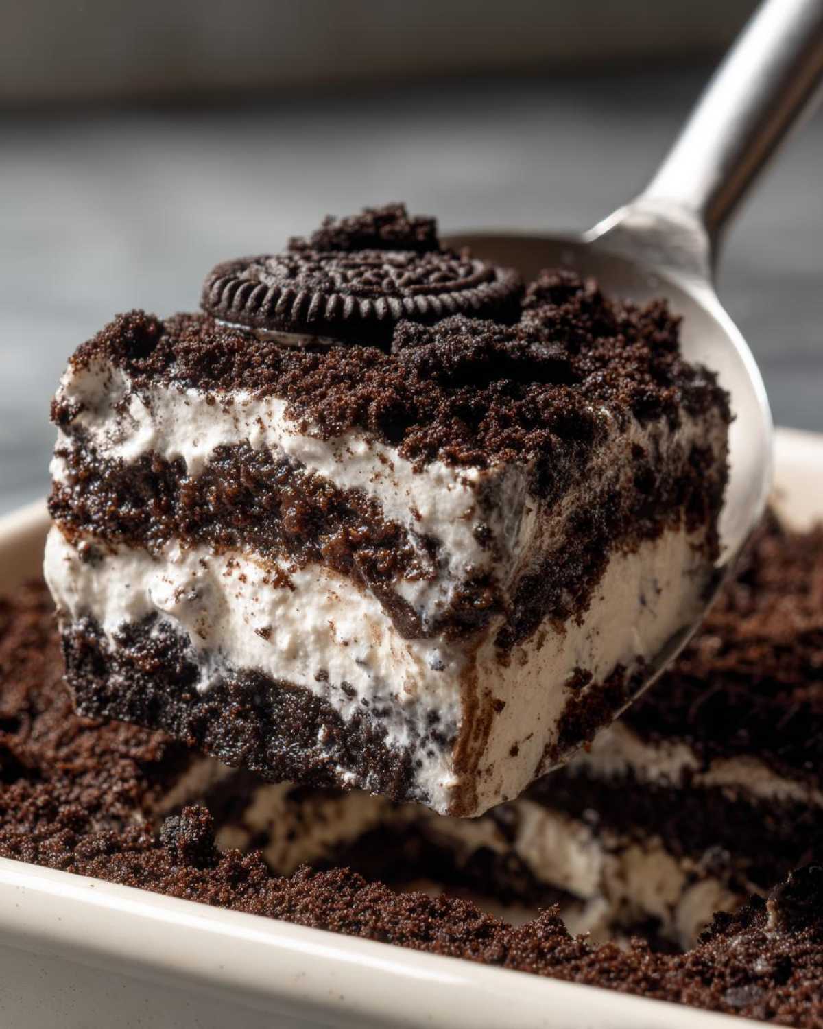 A scoop of creamy, layered dirt cake being lifted out of a dish with a metal spoon, topped with cookie crumbs and a whole cookie.
