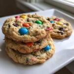 A stack of three freshly baked, chewy M&M cookies loaded with colorful candies, resting on a white plate.