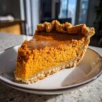 A close-up slice of rich, orange sweet potato pie with a flaky crust, sitting on a light plate.