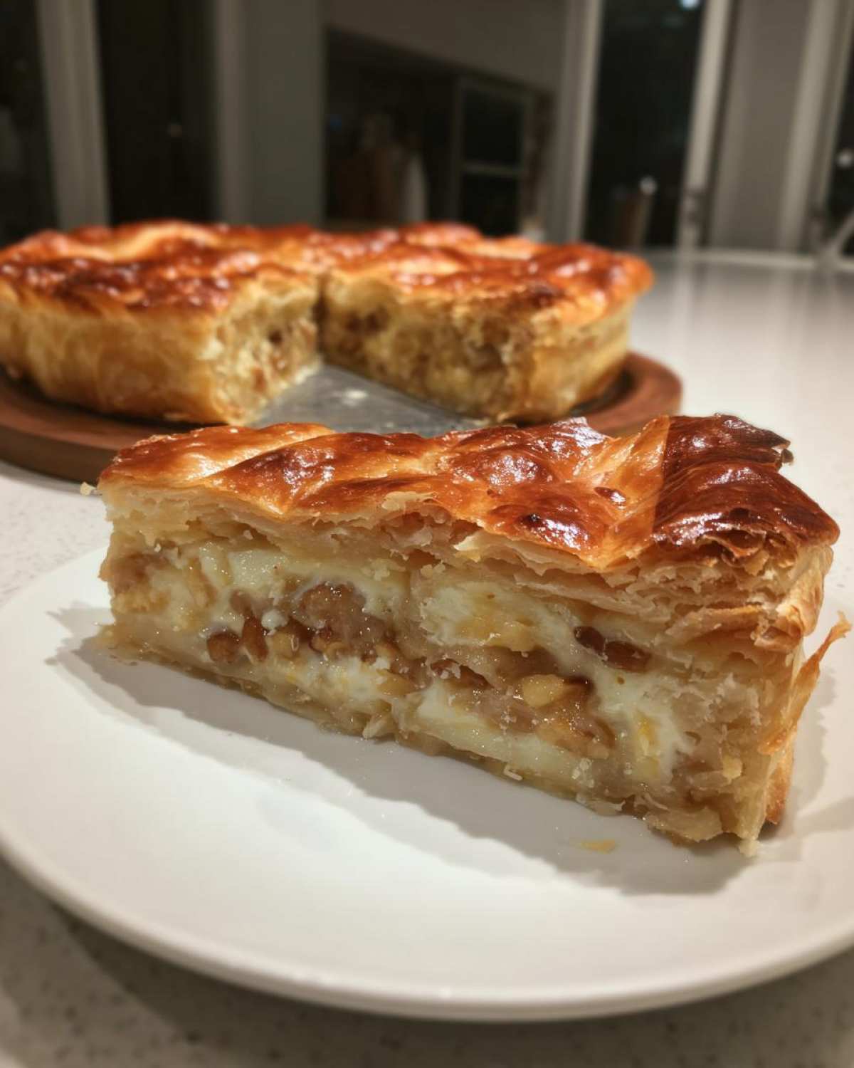 A slice of golden, flaky pastry filled with creamy cheese and nuts, resembling baked brie, served on a white plate.