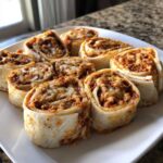 A close-up of several baked taco pinwheels filled with seasoned ground meat and melted cheese, arranged on a white plate.