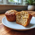 Two banana oatmeal muffins on a white plate, one cut in half showing the moist, textured interior.