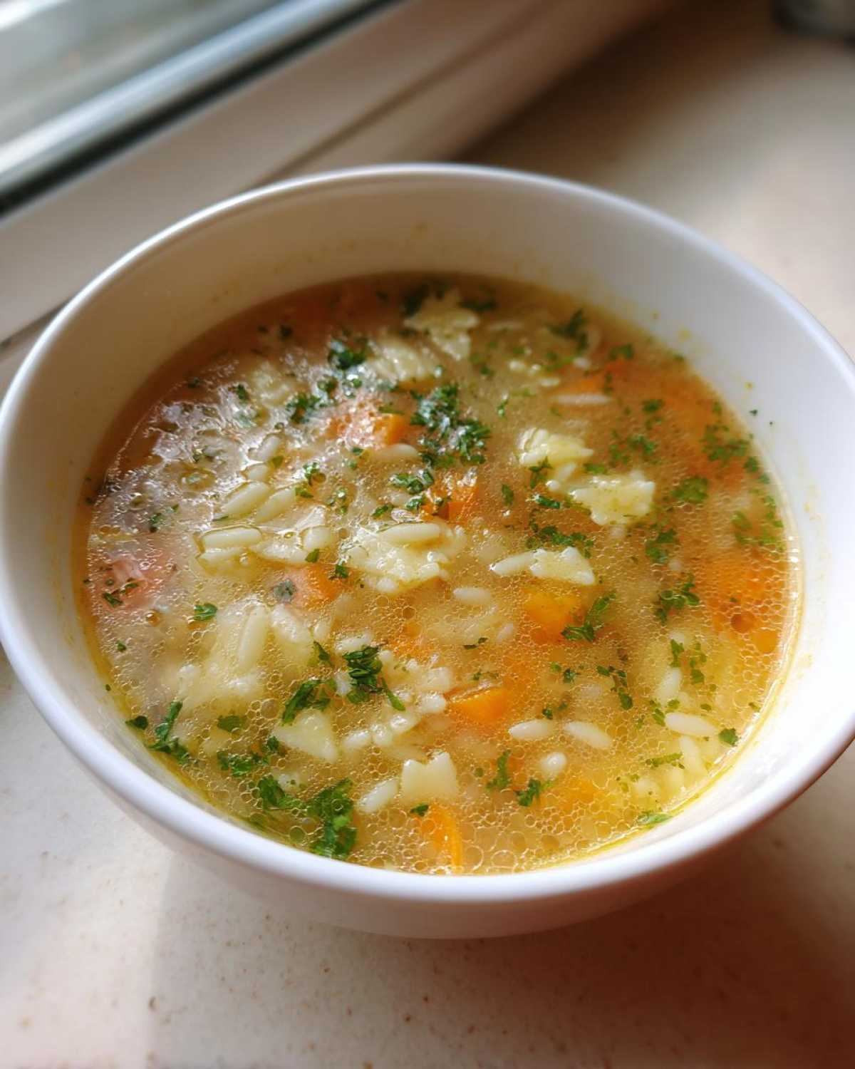 Close-up of a white bowl filled with warm pastina soup, featuring small pasta shapes, diced carrots, and fresh parsley.