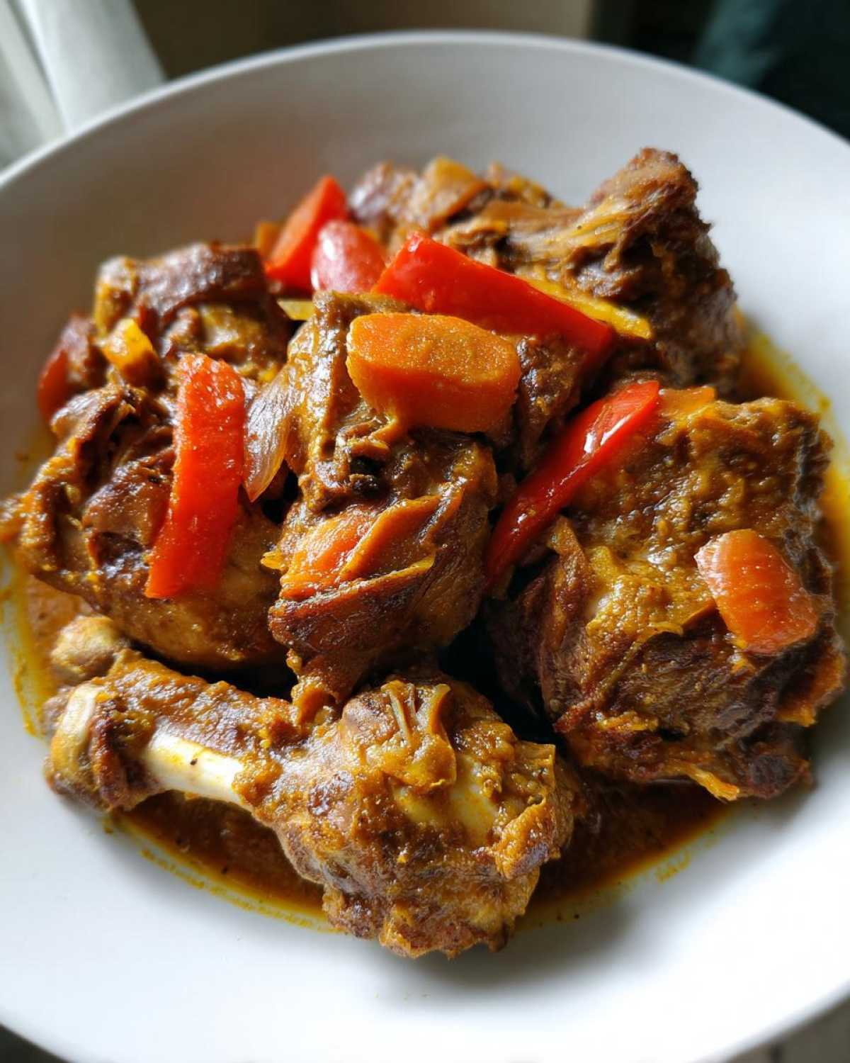 Close-up of tender brown stew chicken pieces served in a white bowl with visible carrots and red peppers.