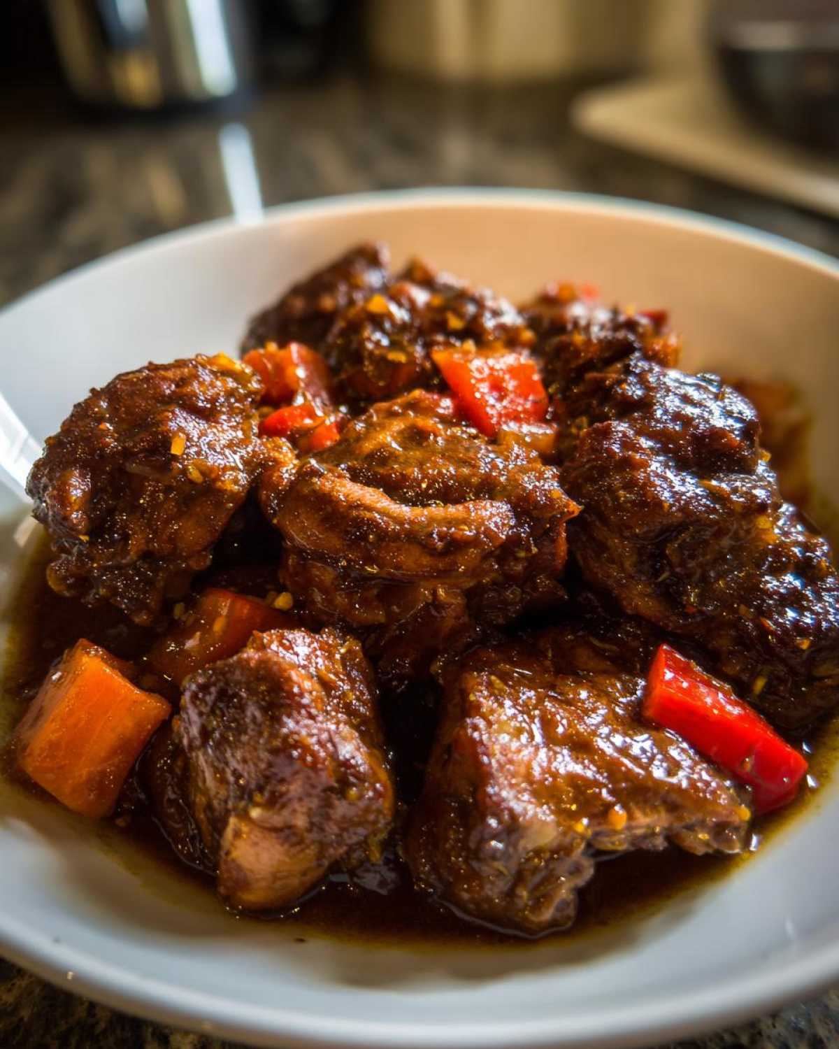 Close-up of tender pieces of brown stew chicken coated in a rich, dark sauce with visible chunks of red pepper.