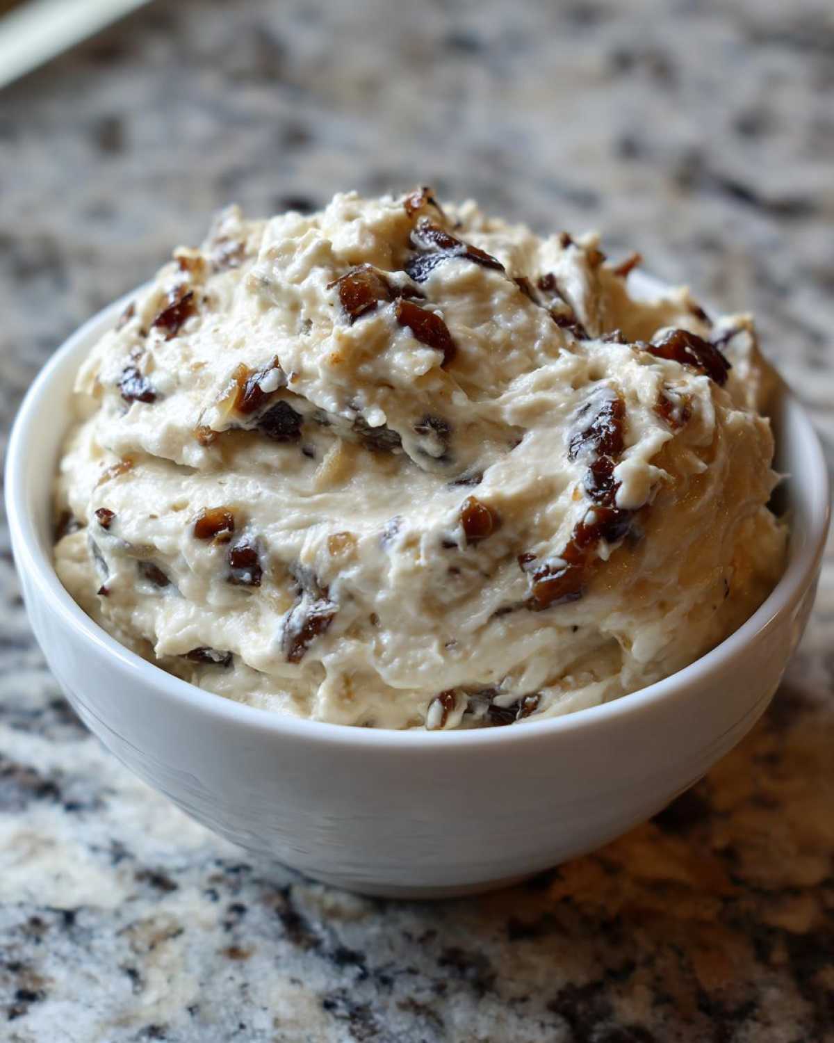Close-up of thick, creamy caramelized onion dip mixed with dark caramelized pieces in a white bowl.