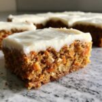A close-up shot of a thick, moist square of carrot cake bars topped with cream cheese frosting.