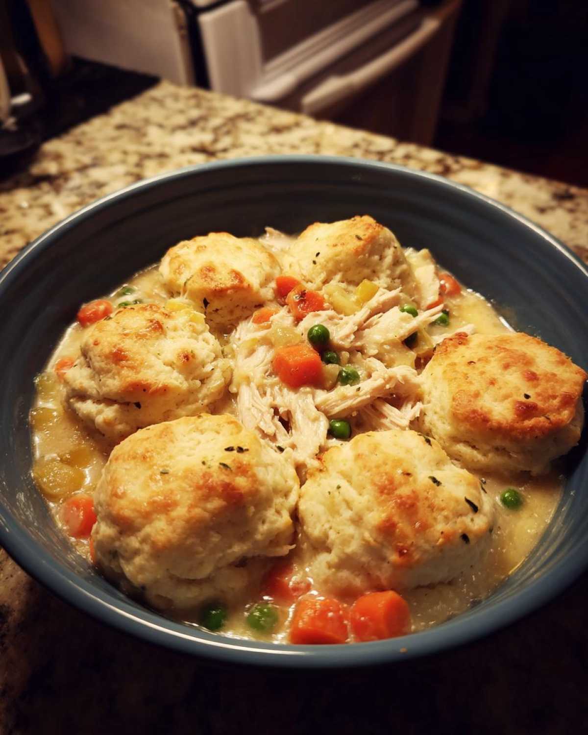 A close-up of a blue bowl filled with creamy chicken and dumplings with biscuits, carrots, and peas.