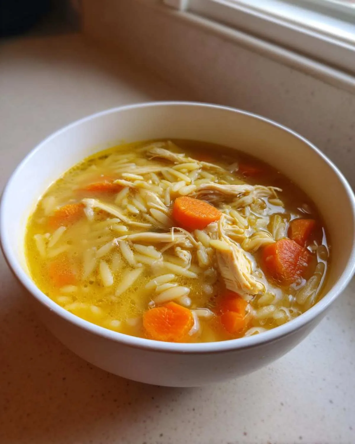 A close-up of a white bowl filled with warm chicken orzo soup, featuring shredded chicken, pasta, and bright orange carrots.