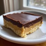 A square slice of rich chocolate peanut butter fudge with a crumbly base, served on a white plate.