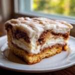 A moist, square slice of cinnamon roll cake with thick cream cheese frosting and a dusting of cinnamon.