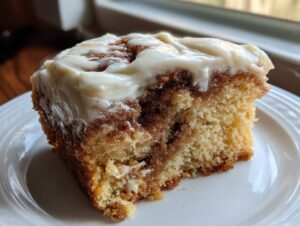 A close-up of a square slice of moist cinnamon roll cake topped with thick cream cheese frosting.