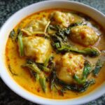 A close-up shot of a white bowl filled with rich, orange coconut curry soup with dumplings and green leafy vegetables.