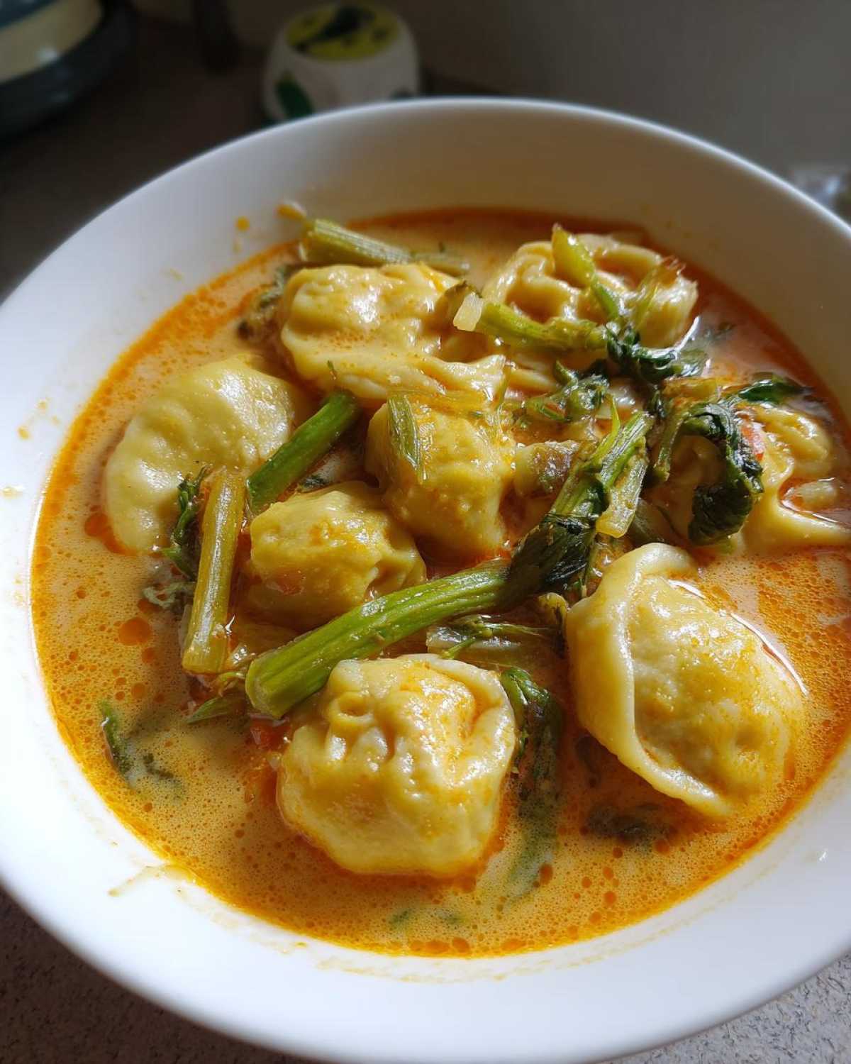 A white bowl filled with rich, orange coconut curry soup featuring several plump dumplings and green vegetables.
