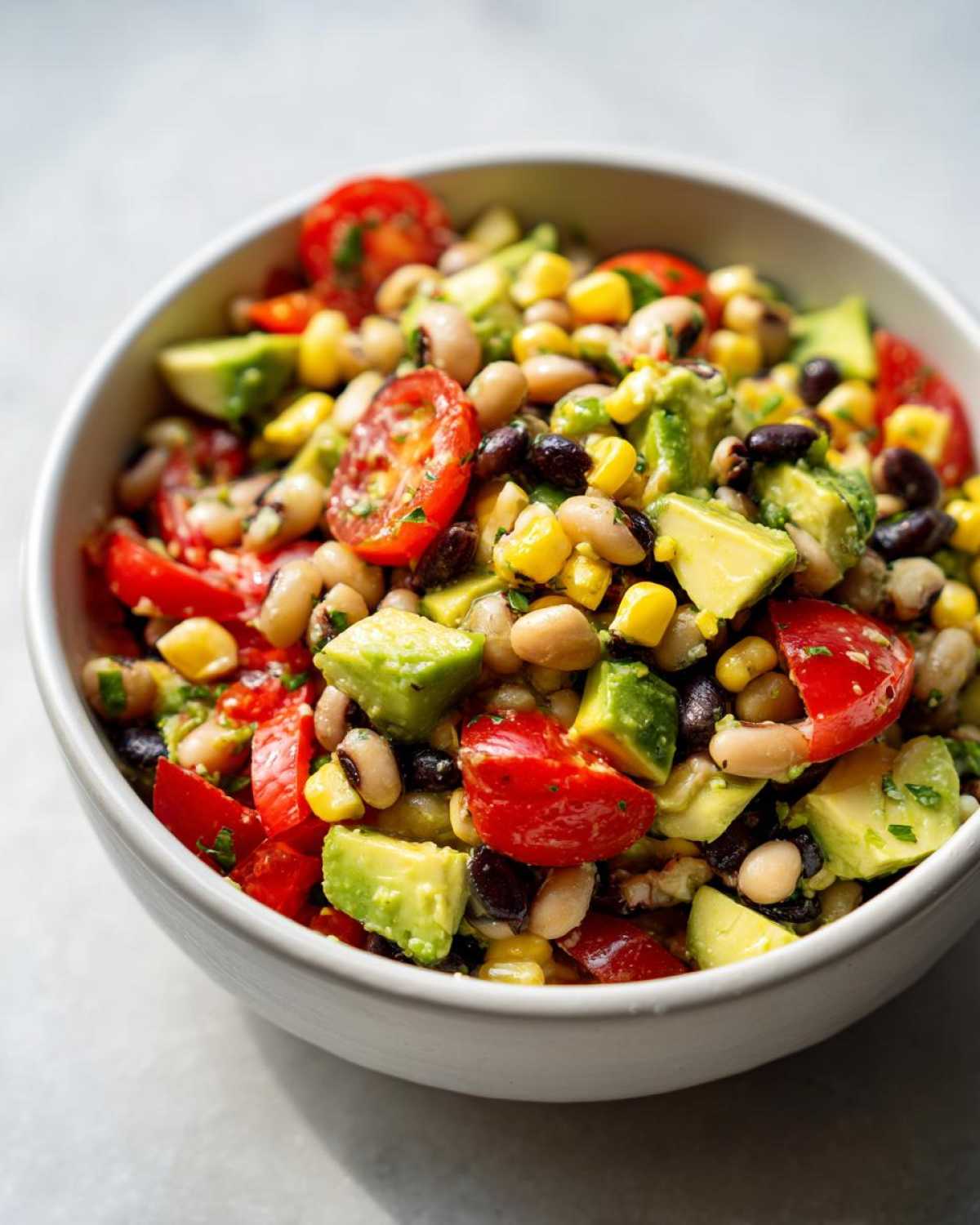 Close-up of a white bowl filled with fresh cowboy caviar recipe ingredients including black beans, black-eyed peas, corn, avocado, and tomatoes.