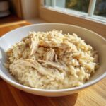 A close-up of creamy crockpot chicken and rice served in a white bowl on a wooden surface.
