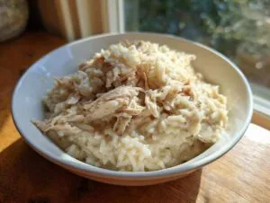 A close-up of creamy crockpot chicken and rice served in a white bowl, highlighted by natural light.