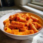 A close-up of a white bowl filled with rich, orange-red creamy tomato pasta made with rigatoni noodles.