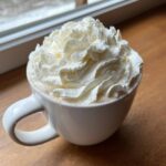 A close-up of a mug filled with creamy white hot chocolate topped high with fresh whipped cream.