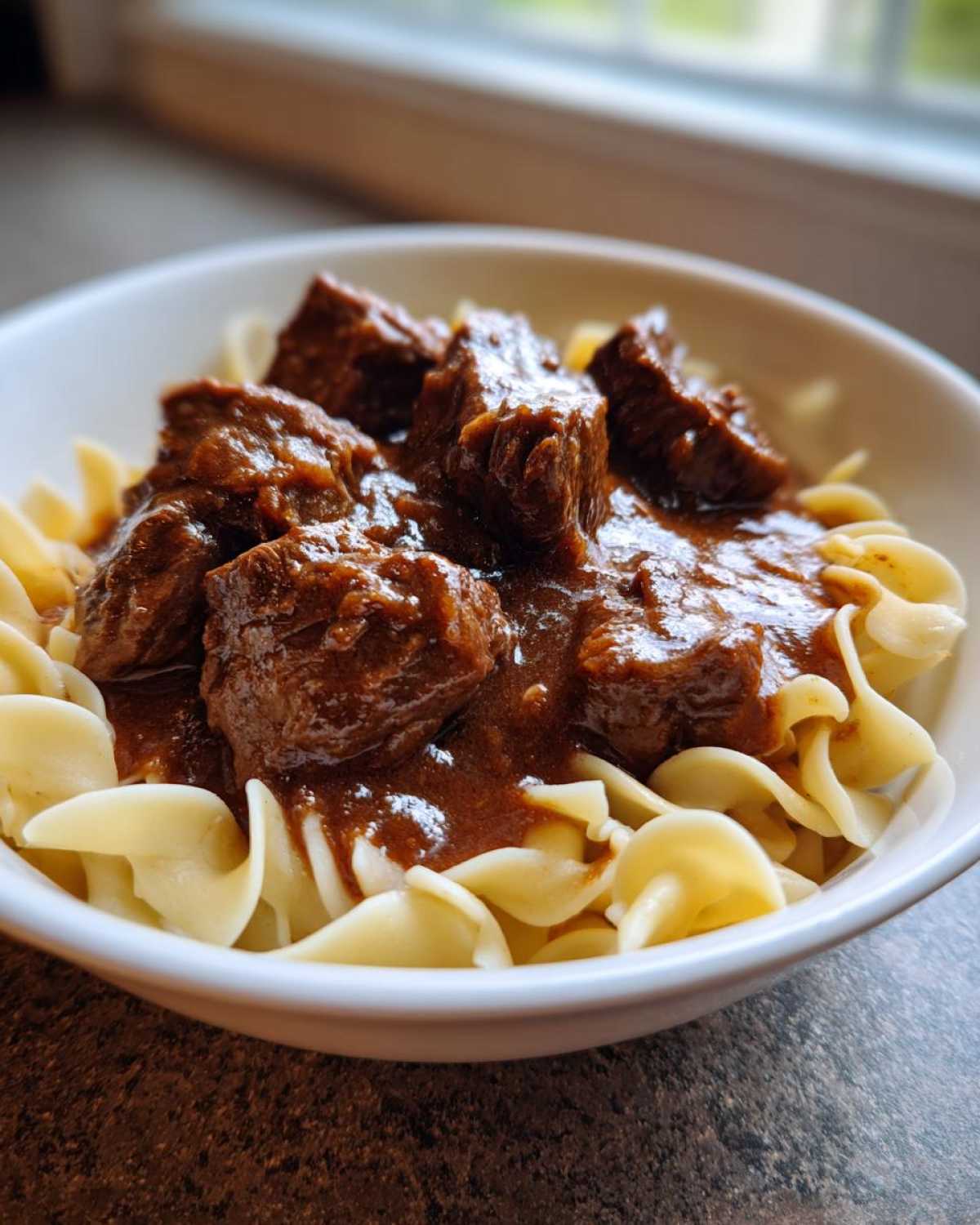 Close-up of tender crockpot beef tips smothered in rich gravy served over egg noodles in a white bowl.