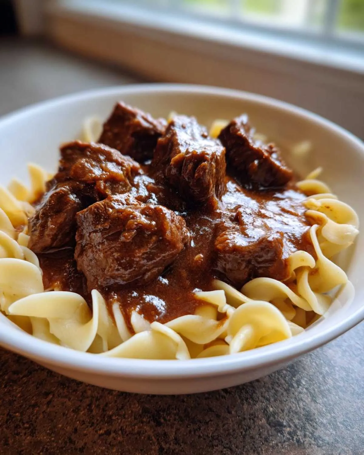 Close-up of tender crockpot beef tips smothered in rich gravy served over egg noodles in a white bowl.