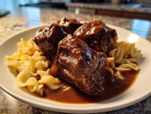 Four tender pieces of crockpot beef tips smothered in rich brown gravy served over egg noodles on a white plate.