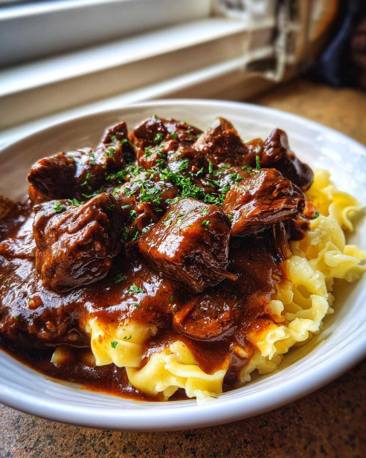 Close-up of tender crockpot beef tips smothered in rich brown gravy served over wide egg noodles and garnished with parsley.
