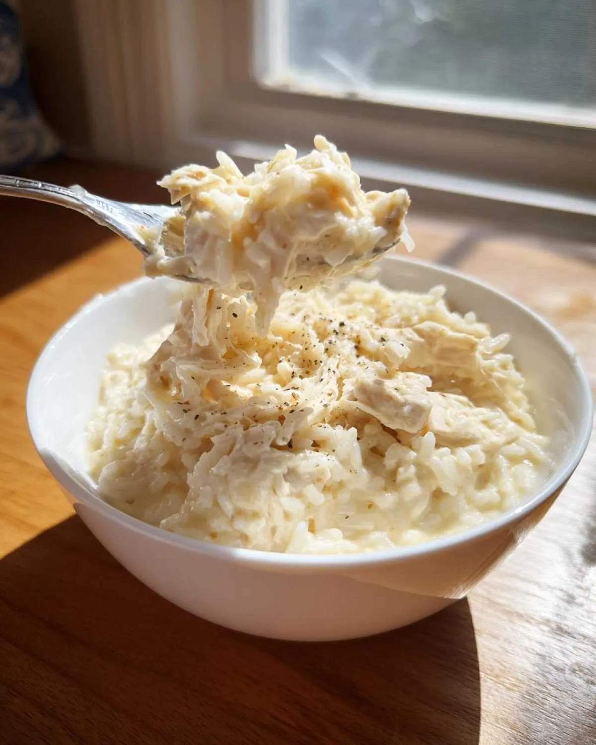 A spoonful of creamy crockpot chicken and rice being lifted from a white bowl, seasoned with pepper.