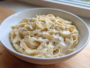 A white bowl filled with fettuccine pasta generously coated in a thick, rich cream sauce and topped with grated cheese.