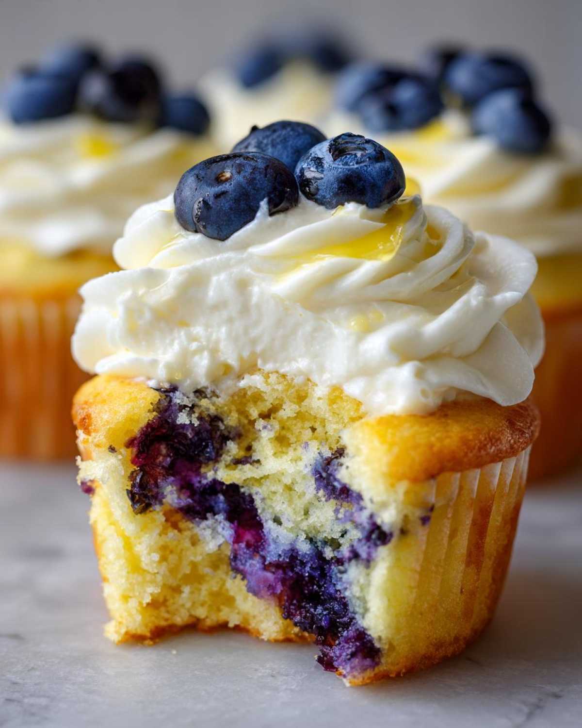 A close-up of a lemon blueberry cupcakes with a bite taken out, showing moist cake and blueberries, topped with white frosting and fresh blueberries.