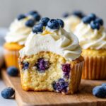 Close-up of a fluffy lemon blueberry cupcakes cut in half showing moist cake and blueberries, topped with cream cheese frosting.