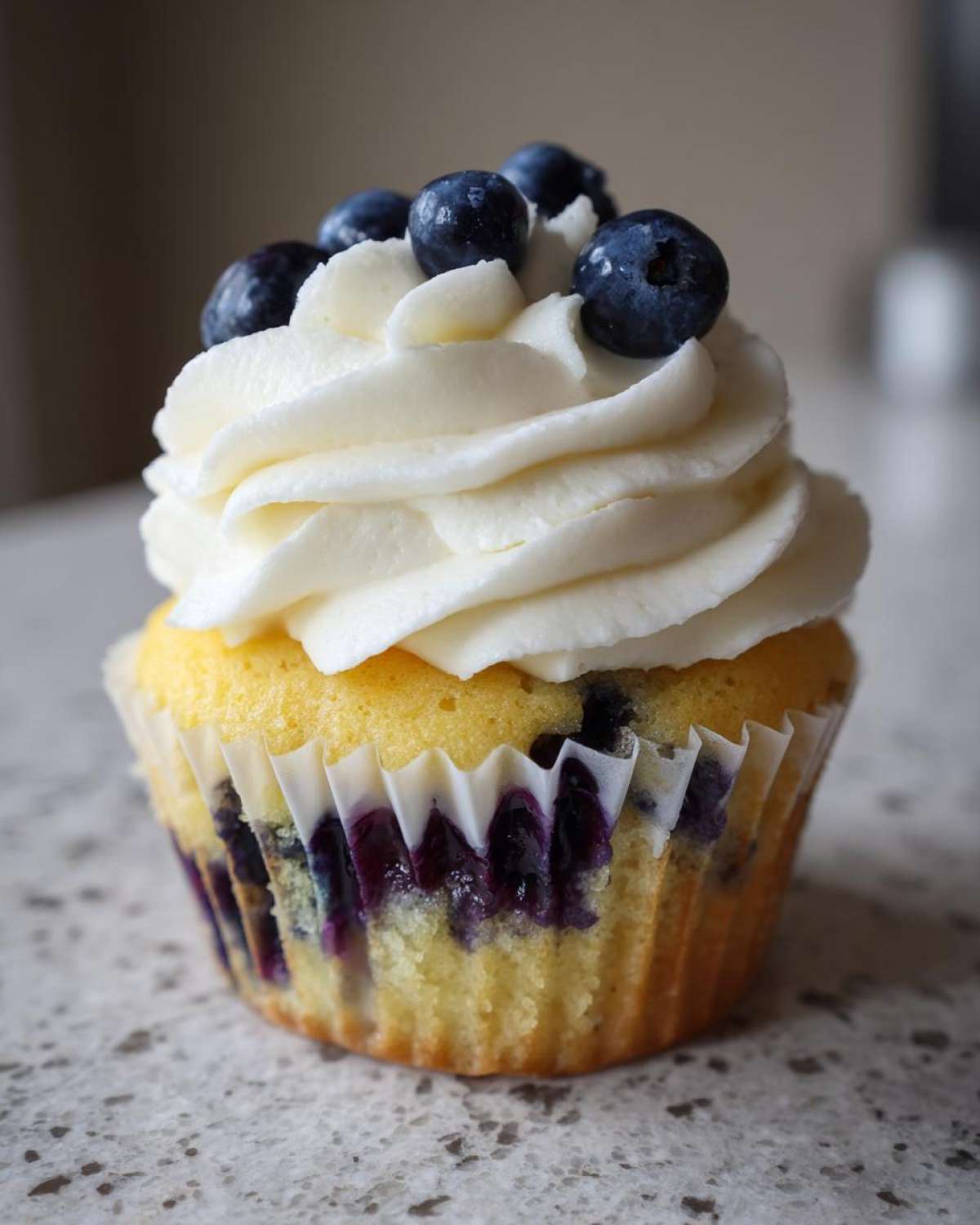 A single, fluffy lemon blueberry cupcake topped with white frosting and fresh blueberries.