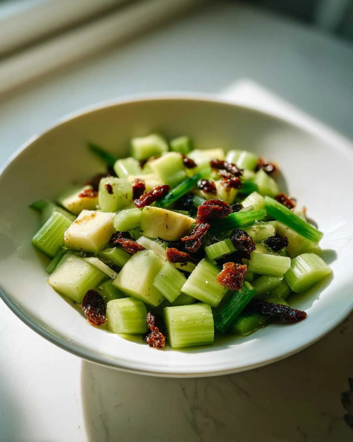 A close-up of a vibrant celery salad featuring chopped celery, apple chunks, and dried cranberries in a white bowl.