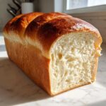 A freshly baked loaf of japanese milk bread, sliced to show its fluffy white interior and shiny golden-brown top.