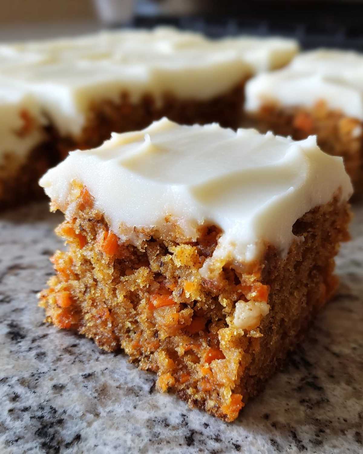 A close-up of a single square of moist carrot cake bars topped with thick cream cheese frosting.
