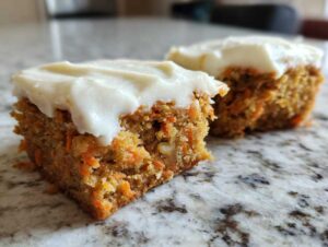 Two squares of moist carrot cake bars topped with thick cream cheese frosting resting on a granite counter.