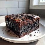 A single, rich, fudgy square of oreo brownies topped with crushed cookies, sitting on a white plate.