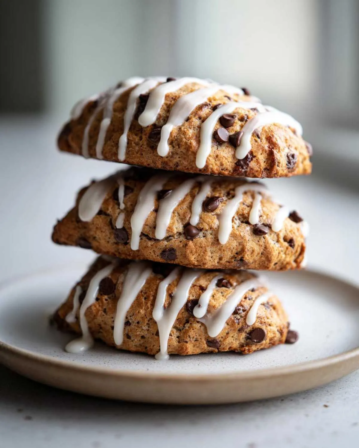 Three delicious chocolate chip scones stacked on a plate, drizzled with white vanilla icing.