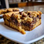 A close-up shot of a thick, gooey chocolate chip cookie bar slice served on a white plate.