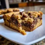 A close-up shot of a thick, gooey chocolate chip cookie bar slice served on a white plate.