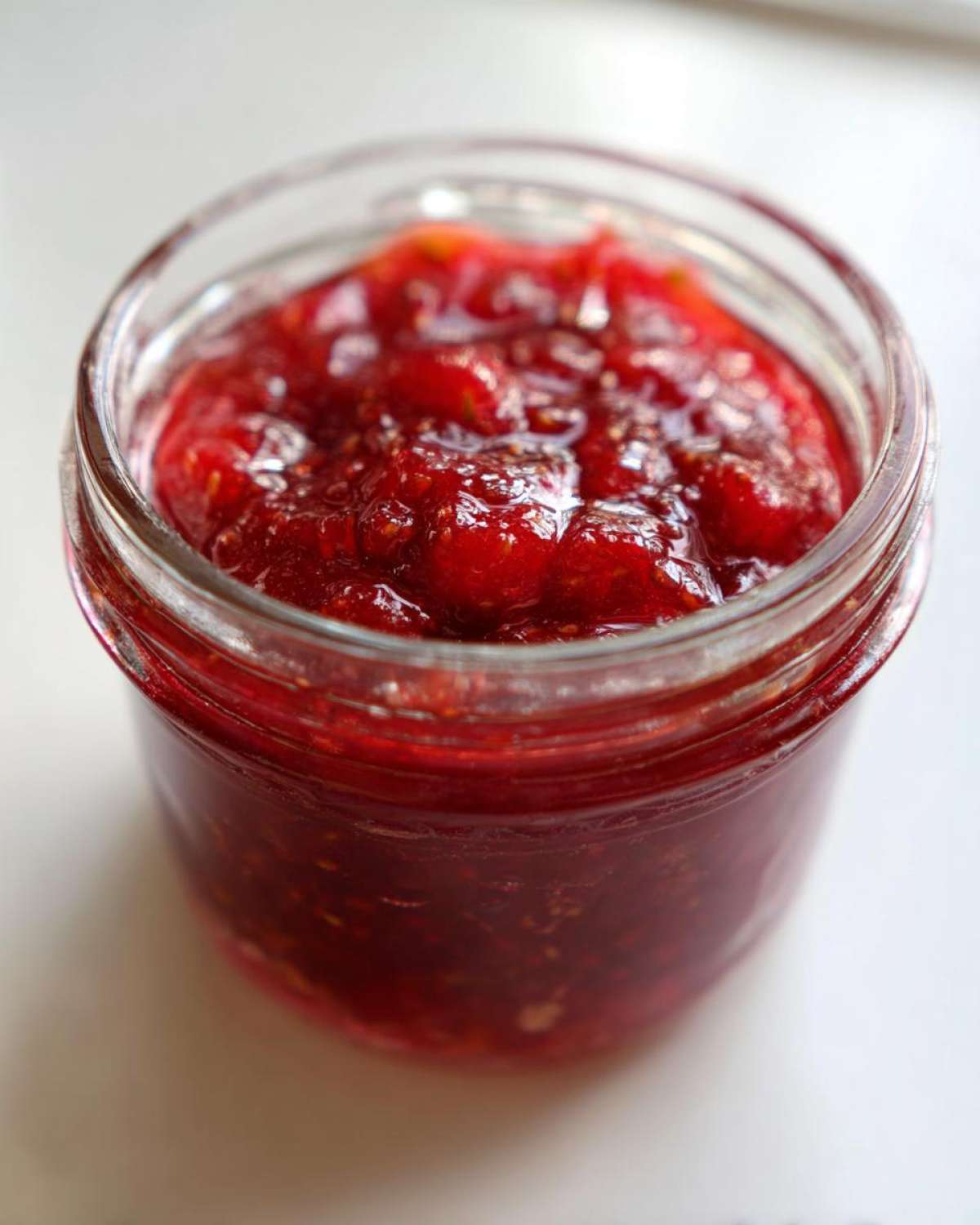 Close-up of a small glass jar filled with bright red, chunky strawberry freezer jam.