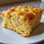 A close-up of a square slice of moist jiffy corn casserole, studded with whole corn kernels, served on a white plate.
