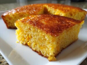 Close-up of a square slice of moist jiffy corn casserole showing whole corn kernels inside and a shiny, golden-brown crust.