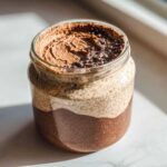 Close-up of layered chocolate chia pudding in a glass jar, topped with chocolate cream and sprinkles.