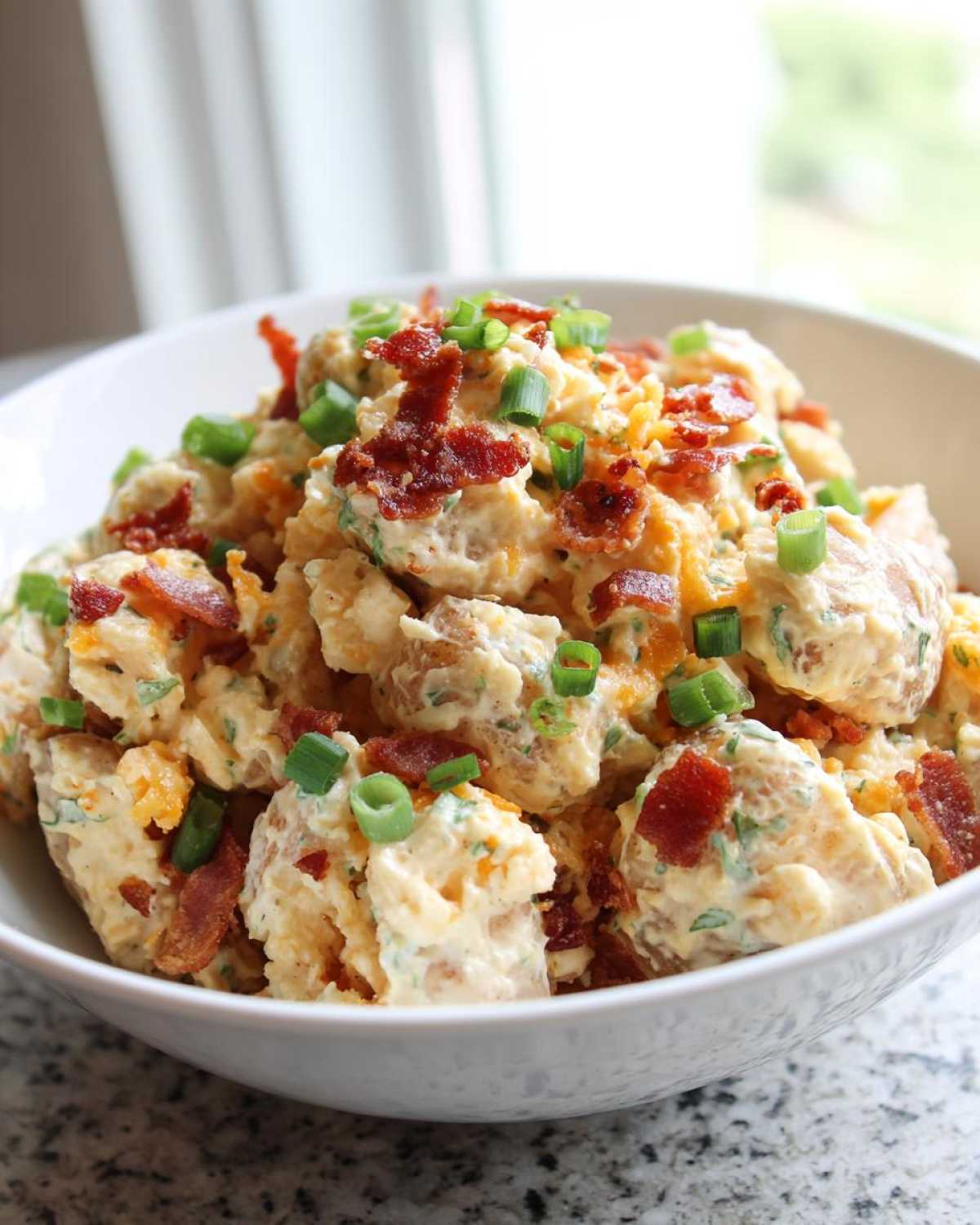 A close-up of loaded baked potato salad topped with bacon bits and green onions in a white bowl.