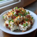 A creamy bowl of loaded baked potato salad topped with bacon bits, shredded cheddar cheese, and green onions.
