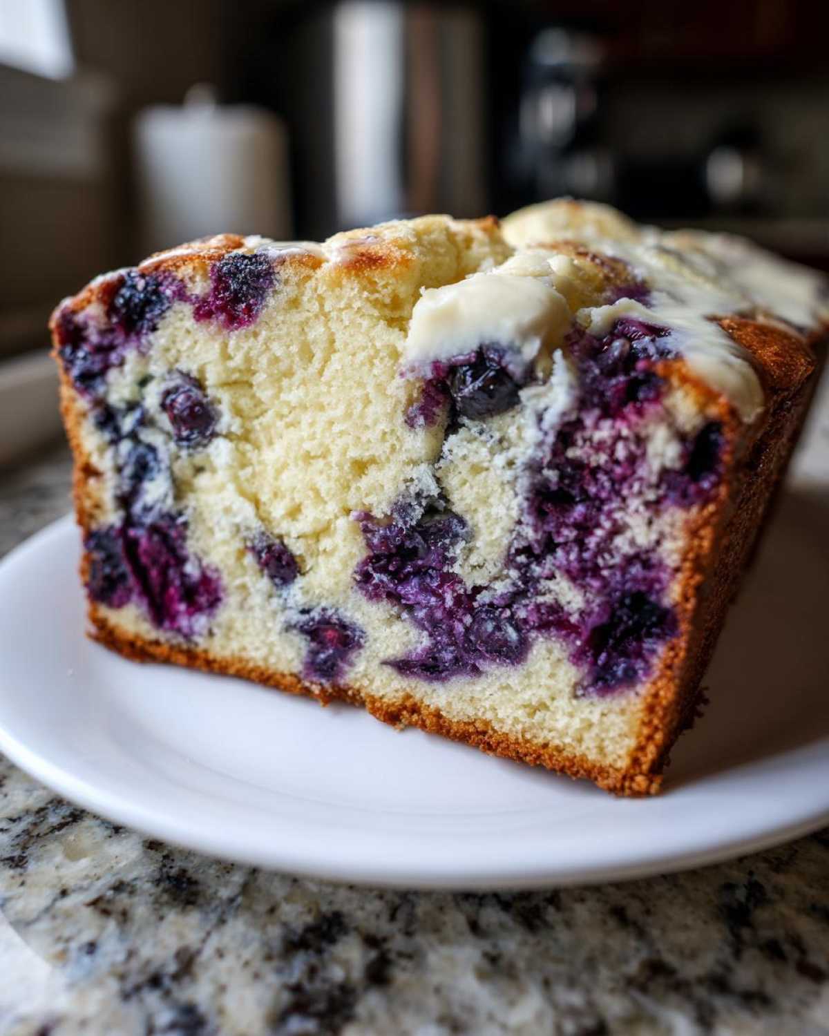 Close-up of a moist slice of blueberry bread showing vibrant purple blueberries baked into the light yellow crumb, topped with a light glaze.